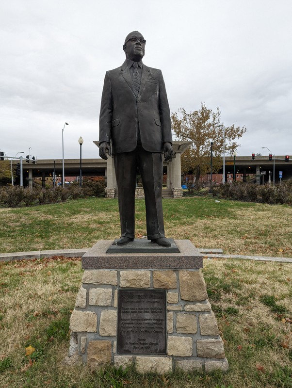 A portrait-oriented picture of a dark-colored metal statue. The statue is a life-sized depiction of an older man in a suit and tie with glasses. The statue is standing on a stone brick plinth with a plaque on the front of it. The plaque is made of the same metal as the statue. Behind the statue, a grassy area can be seen, with a concrete pergola surrounded by bushes and a small tree. Further in the background, a highway overpass can be seen.