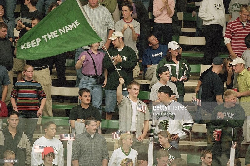 Men and a few women stand to cheer on the team. A green sweatshirt and flag feature the "Sioux" mascot.