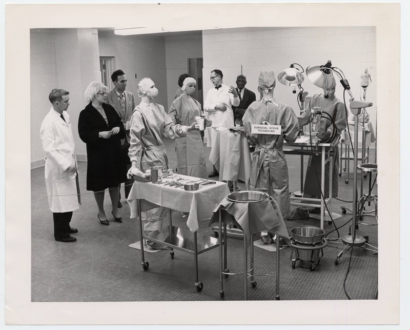 Black and white photograph of a physician giving a demonstration of a surgical team operating environment to a crowd of people. View includes models, staged demonstration area and a "Surgical Scrub Technician."