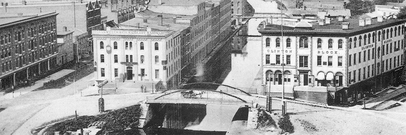 This artist depiction (date unknown) shows the Erie Canal in the center with the Amos Building to its right. The building used to sit along the banks of the Erie Canal, but that is now Erie Blvd.  