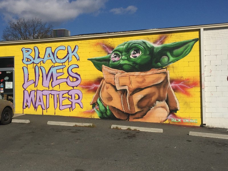 A blue and purple script of Black Lives Matter is painted on the wall next to a mural of Baby Yoda who is green which a brown cloak.
