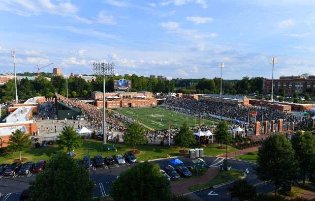 A wide angle shot of the stadium during a football game in the late afternoon. The parking lot and football field can be seen. 