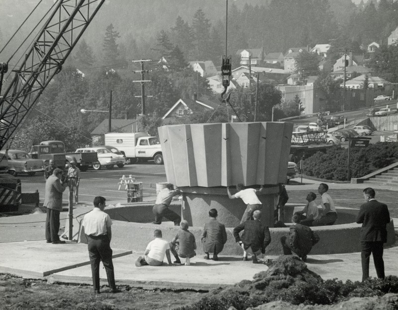 Black and white photograph depicts a crowd of people watching a large round modernist object being placed on a round cement slab. A parking lot, houses and treed hills are visible in the background.