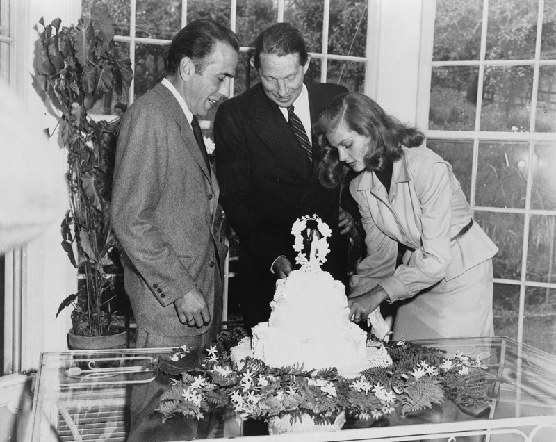 Black and white photo of two men and a woman. The woman is cutting the wedding cake in the center of the table while the two men observe her to the left.