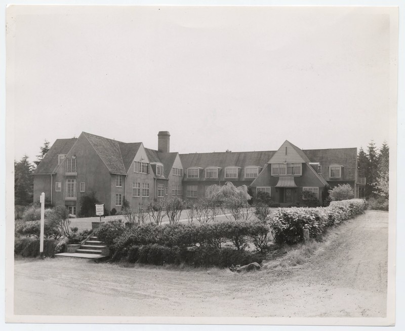 Black and white photograph of an exterior view of the Nurses Dormitory on the Marquam Hill campus, Gaines Hall. The storybook style building is framed by a lawn and shrubbery. A small sign reads "University of Oregon Medical School. Gaines Hall."