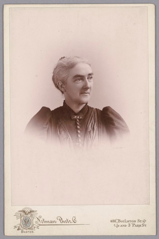 Black & white photo of a woman, grey hair pulled back into a bun, in a dark high-necked blouse with puffed sleeves.