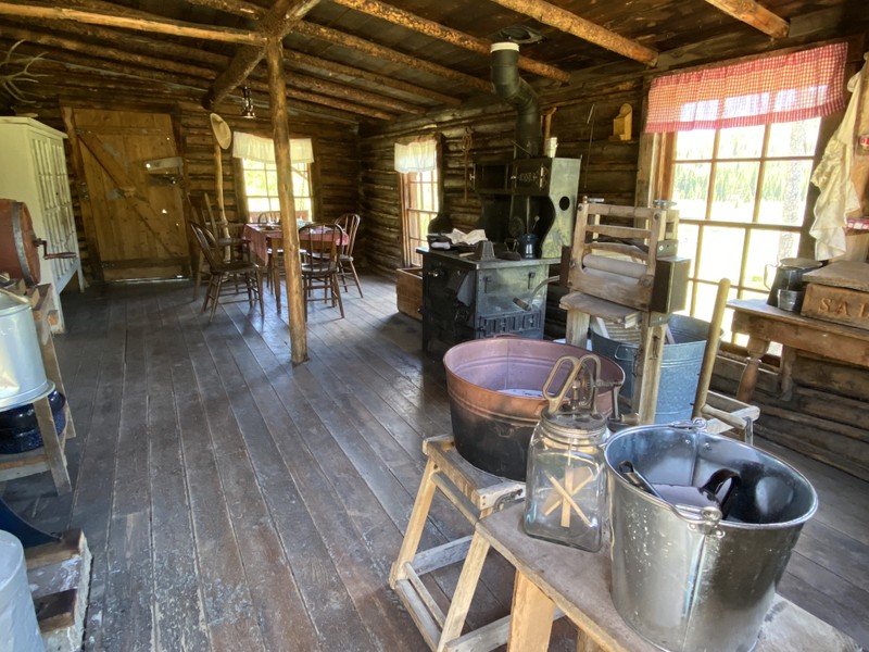 The interior of the cabin, showing a Daisy butter churner, a washing bin, a wood-burning stove, and a set dining table