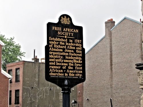 Black sign with gold text, photographed at an angle. Brick buildings are visible in the background.