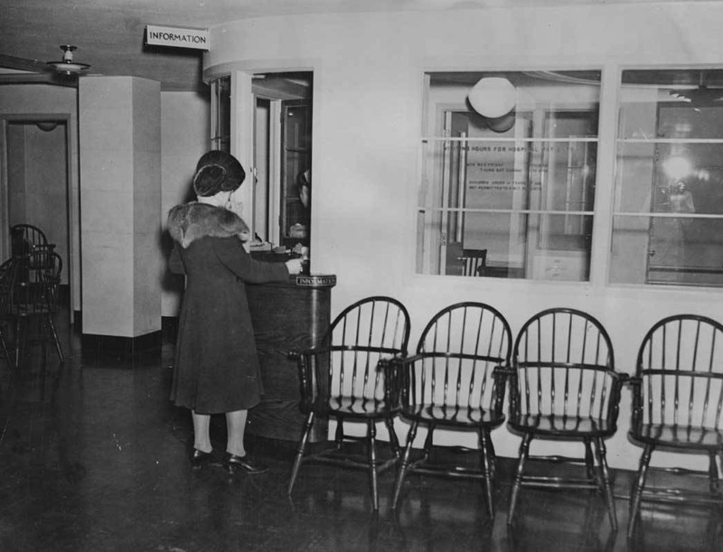 A woman is viewed from behind standing at a small admitting window in a waiting room filled with chairs.