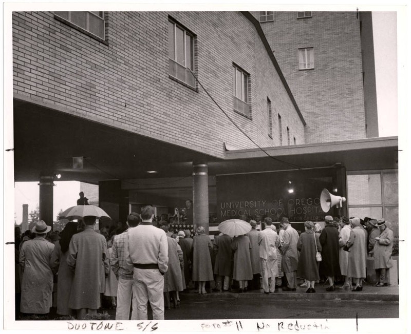 Black and white photograph of the dedication celebration of the University of Oregon Medical School Hospital. A crowd of onlookers watch several figures at the top of stairs underneath a pedestrian bridge connecting two buildings.