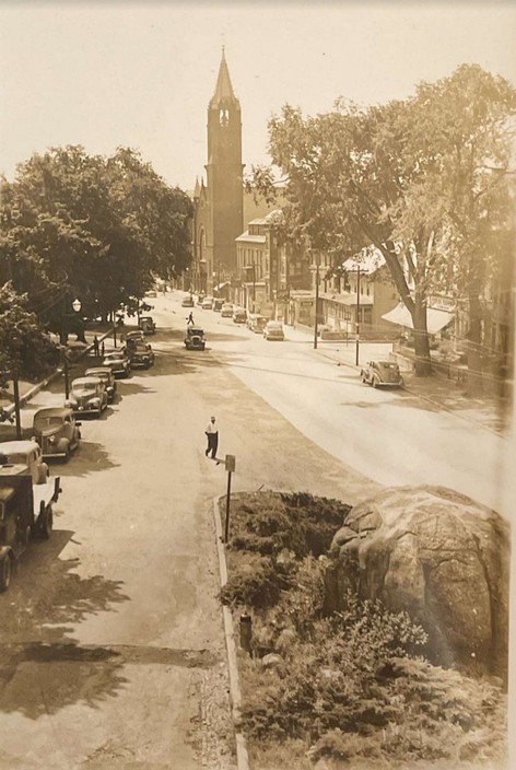 A black and white photograph of a street with the rollstone boulder sitting in a grassy area in the middle of the street.  