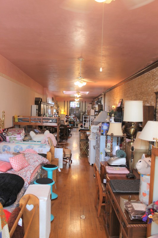 A narrow, deep showroom space crowded with furniture for sale. The walls are made of sand colored brick.