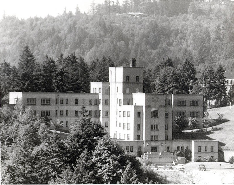 A large white building is viewed among hills covered with large evergreen trees.