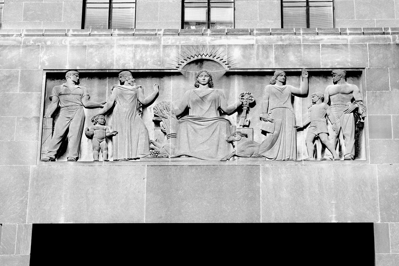 An image of the central panel of the 6th Floor Frieze on the South Face of City Hall. Center: A symbolic female figure representing Kansas City, seated, holding a sheaf of grain and the key to the city, surrounded by symbolic figures representing civic virtues. Left: Three figures representing Education with Law as its guardian. Right: Three figures, Faith holding the scroll of the future and Public Spirit represented by a young man and a boy.