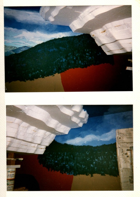 These photos show the early stages of creating the rock shelter. The white foamcore is attached to the ceiling and walls in the corner of the room. The edges are curved to help with the shape of the rock shelter later in the process. The walls are blue with the sky in the mural and unfinished trees sit on the hills surrounding the rock shelter on the walls. 