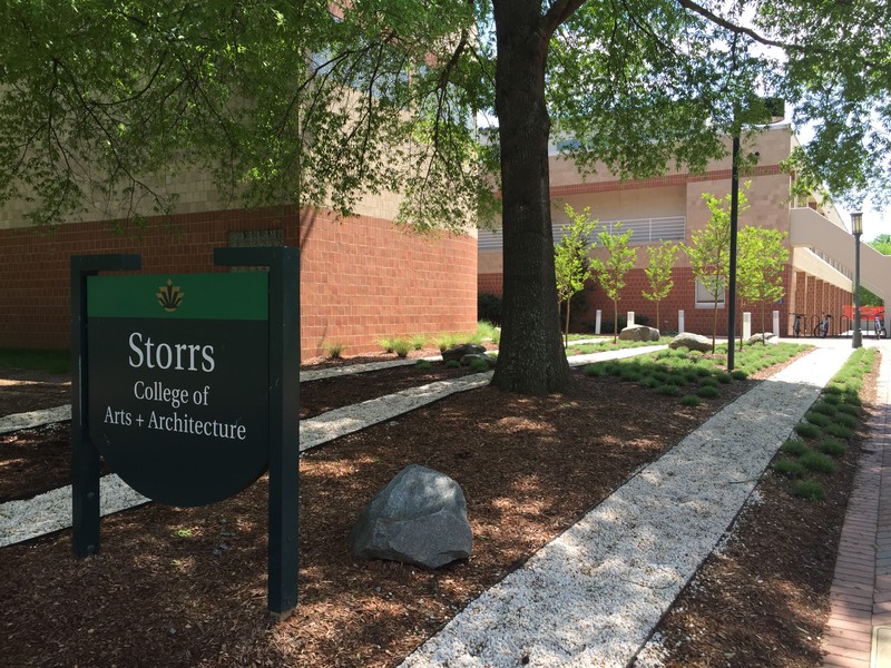 Black sign with white lettering marking the location of Storrs Hall, UNC Charlotte, 2015. Storrs Hall and a tree are also in view.