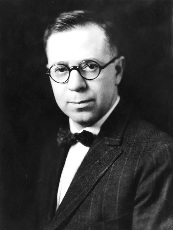 A black and white photo shows a man in a dark striped suit and bowtie. He wears wire-rimmed glasses.