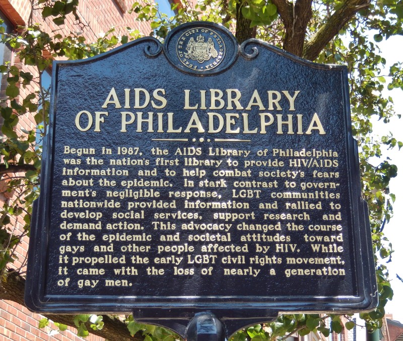 Black sign with gold lettering, reading "Begun in 1987, the AIDS Library of Philadelphia was the nation's first library to provide HIV/AIDS information and to help combat society's fears about the epidemic. In stark contrast to government's negligible response, LGBT communities nationwide provided information and rallied to develop social services, support research and demand action. This advocacy changed the course of the epidemic and societal attitudes toward gays and other people affected by HIV. While it propelled the early LGBT civil rights movement, it came with the loss of nearly a generation of gay men."