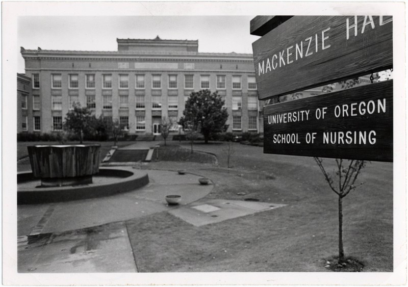 Black and white photograph shows sign in the foreground: Mackenzie Hall, University of Oregon School of Nursing. Behind the sign stands a round fountain plaza. Behind that, a large light colored brick building stands. 