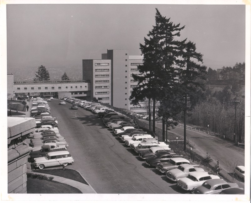 Black and white photograph of the University of Oregon Medical School Hospital, viewed from the west, and the skybridge connecting to the Outpatient Clinic. A surface parking lot is visible in the foreground.