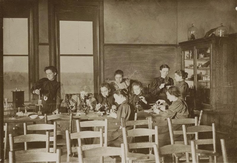 Black & White Photo. Classroom chairs & tables scattered, women in 19th century clothing