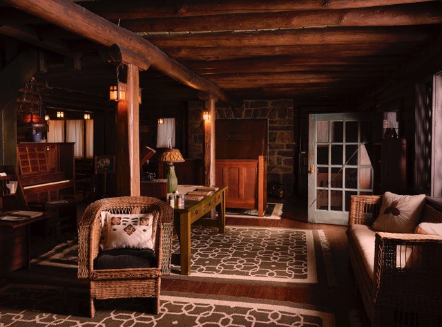 An interior view of the Log House at Craftsman Farms showing the living room, with log walls and beams. In the foreground, in the center of the large room is a willow chair with an embroidered pillow, and other craftsman style wood furnishings throughout the room. A large stone fireplace with a copper hood is seen at the far end of the room and to the right an open door lets in some sunlight.