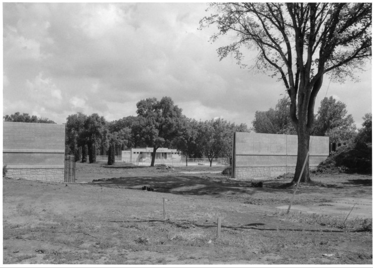 Black and white photo of bath house building surrounded by trees. Seen through opening in brick floodwall.