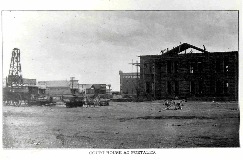 Photo of the original courthouse being constructed. Four men on top of courthouse working on the building. Other buildings and businesses also pictured. 