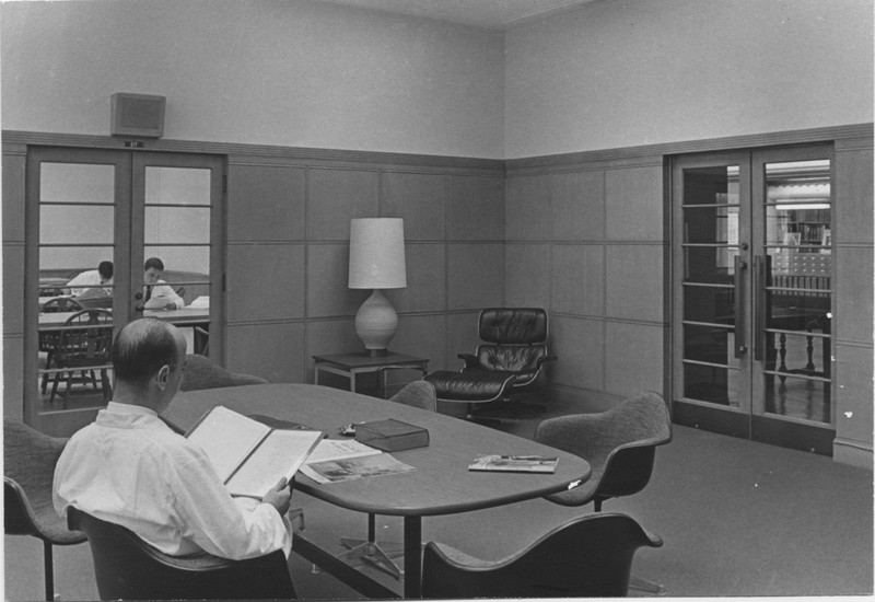 Black and white photograph of an unidentified man reading in the Physicians Reading room located in the Old Library. In the corner, an Eames-style chair is visible next to a lamp.