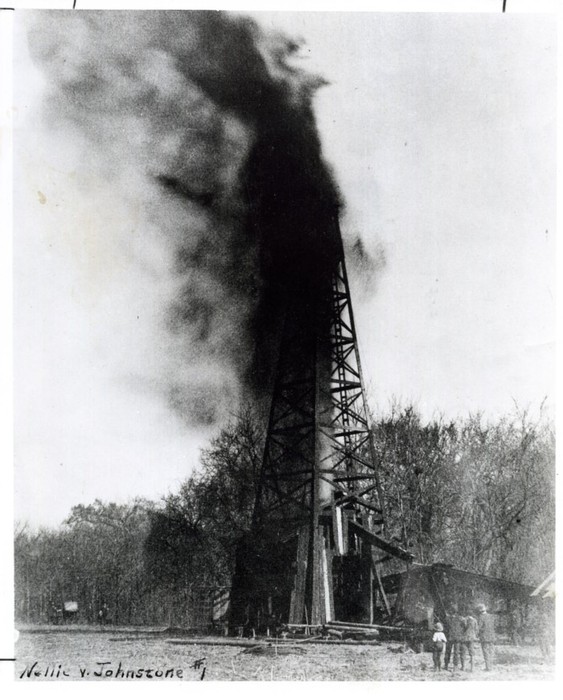 A black and white photo of oil shooting up over an oil derrick.