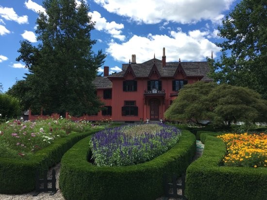 An exterior view of Roseland Cottage and its garden. Circular hedges surround a garden of purple flowers. In the background, the red Roseland Cottage is visible.