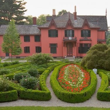 An exterior view of Roseland Cottage and its garden. Circular hedges surround a garden of multicolored flowers. In the background, the red Roseland Cottage is visible.