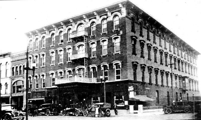 A black and white photo of a street scene in the early 1920's. A row of buildings is seen. Second from left is the C.H. Little Building. A number of period cars and bustling shoppers are seen. 