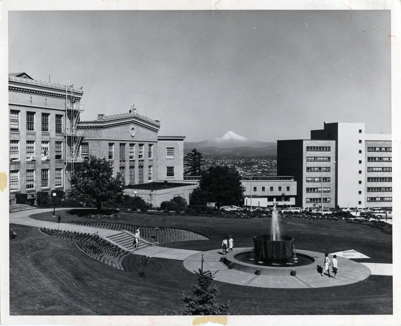 Black and white photograph of Mackenzie Hall, Baird Hall, pedestrian bridge and Medical School Hospital with Mount Hood in the background and Alumni Fountain in the foreground.