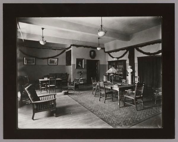  Black & White photo of a table with a tiffany lamp, chairs. Bookcases and doorways in the background.