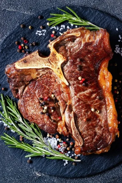 Gebratenes Porterhouse Steak auf dunklem Holzuntergrund, angerichtet mit grünem Pfeffer, Rosmarin und Salzflocken Gebratenes Porterhouse Steak auf dunklem Holzuntergrund, angerichtet mit grünem Pfeffer, Rosmarin und Salzflocken