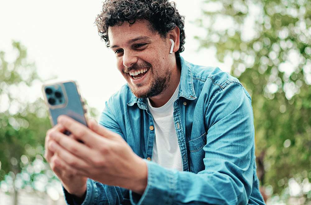 Fundo com árvores. Homem sentado olhando o celular, segurando-o com as duas mãoes e usando um fone de ouvido na orelha