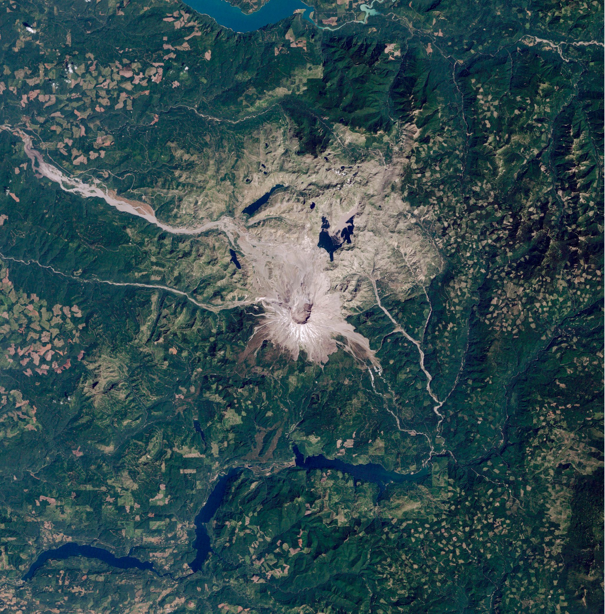 Mittig im Bild ist der grau-braune Mount St. Helens zu sehen sowie die ihn umgebende, gleichfarbige Vulkanlandschaft. Die Satellitenaufnahme zeigt um das Gebiet herum grüne Landschaft und oben und unten im Bild blaue Stauseen.