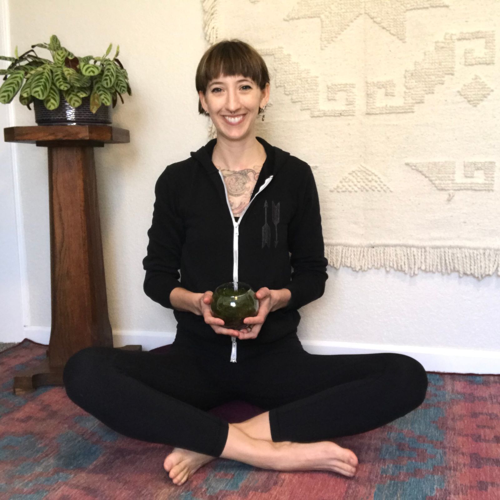Becky, a white genderqueer person, sits cross-legged on a cozy rug in front of a wall with a Mexican blanket. There is a plant on plant stand next to them. They are wearing a black hoodie and leggings, and smiling while holding a small moss terrarium. 