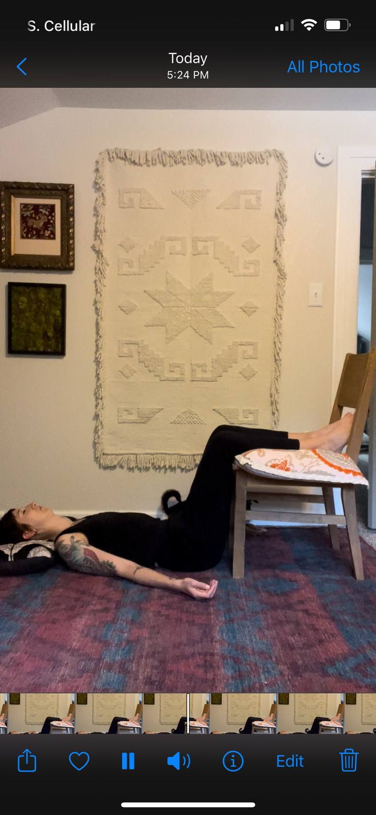 Screen shot of a video still of Becky lying on a colorful rug in "legs up the chair" pose. Becky is a white non-binary person in black tank top and pants. Their head is on a pillow and their lower legs are resting on the seat of a cushioned chair.