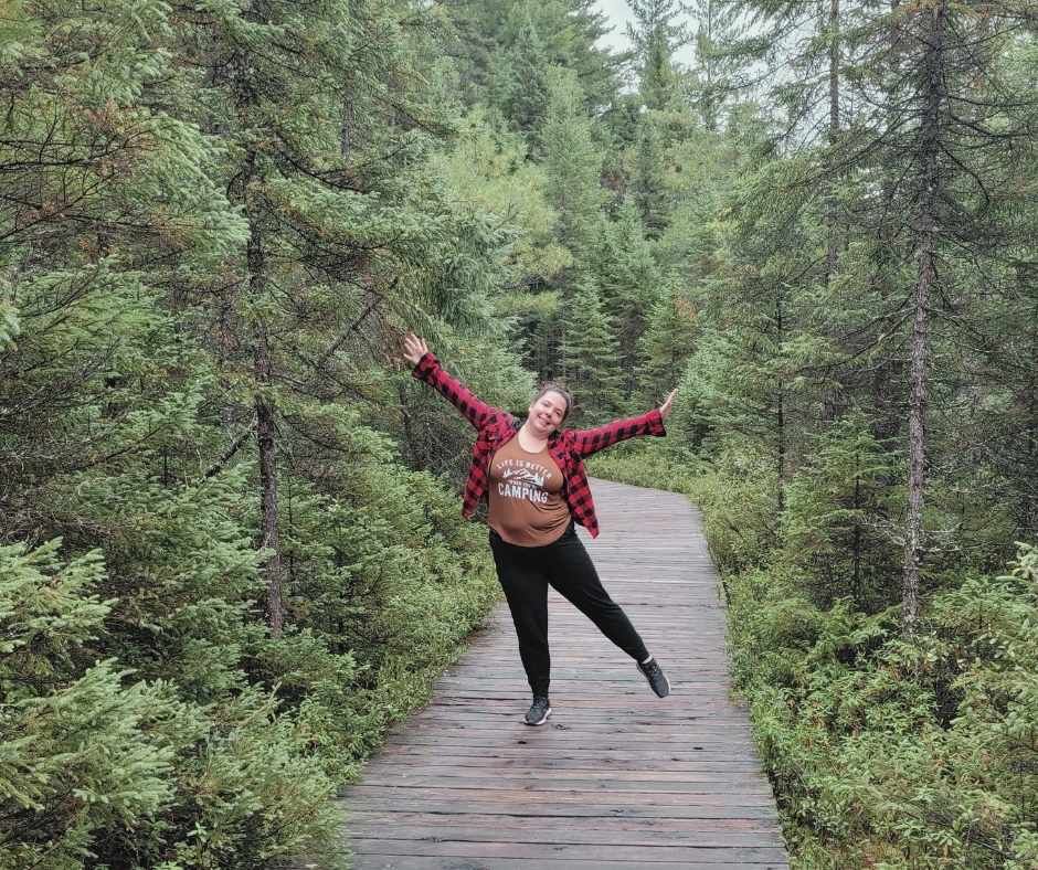 Billie stands on a boardwalk surrounded by trees.  Billie is standing on one foot with arms and leg on right out.  Billie is smiling, looking tired. Wearing black pants, sneakers, a brown tank that says 'I'd rather be camping' and a plaid shirt on top.