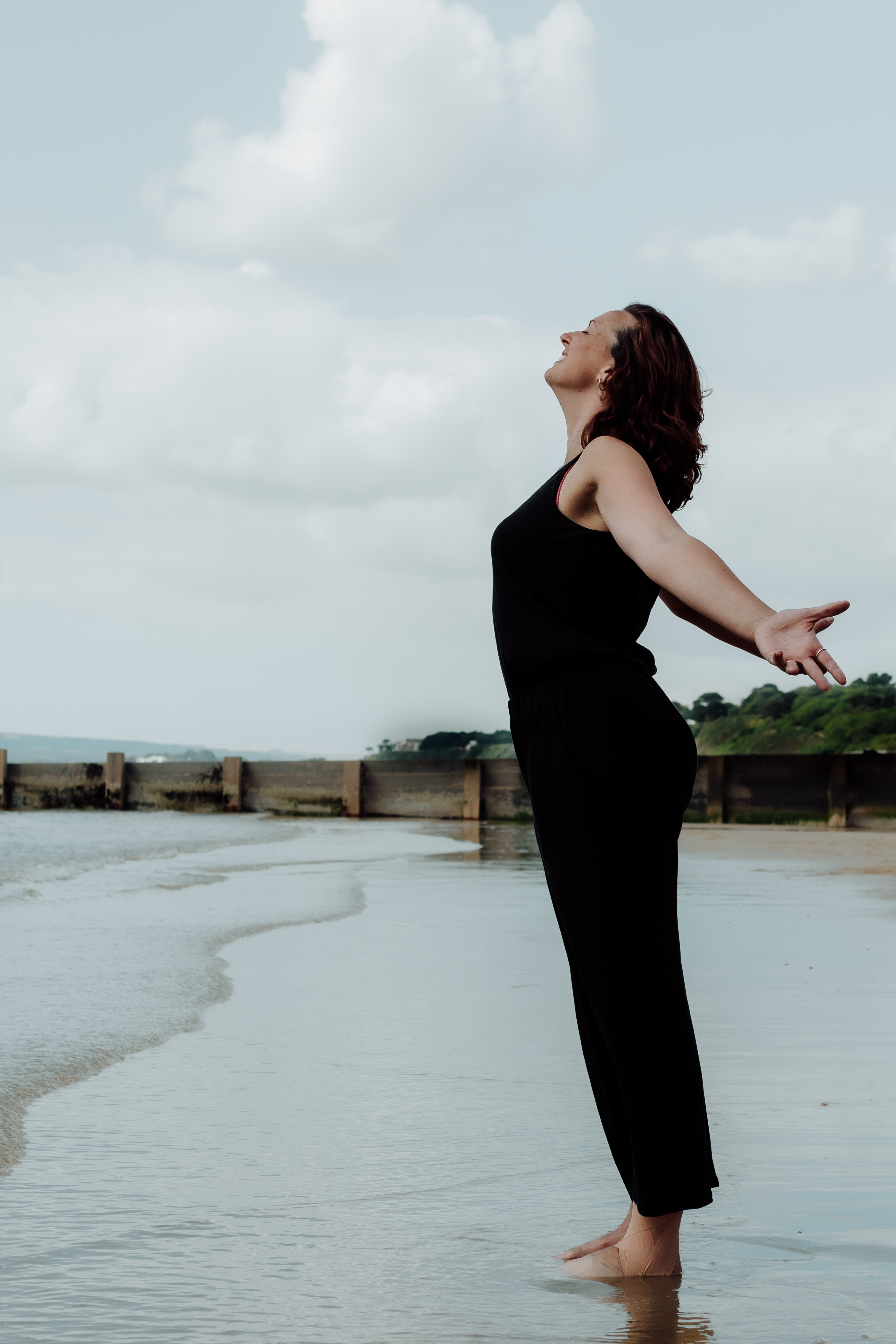 Emma, a yoga teacher and holistic health coach with arms outstretched and heart towards the sky on the beach by the shore