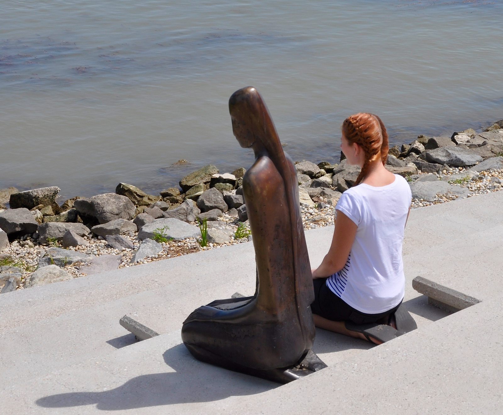 A woman kneeling beside a sculpture of a kneeling woman, both facing a river, symbolizing reflection and stillness.