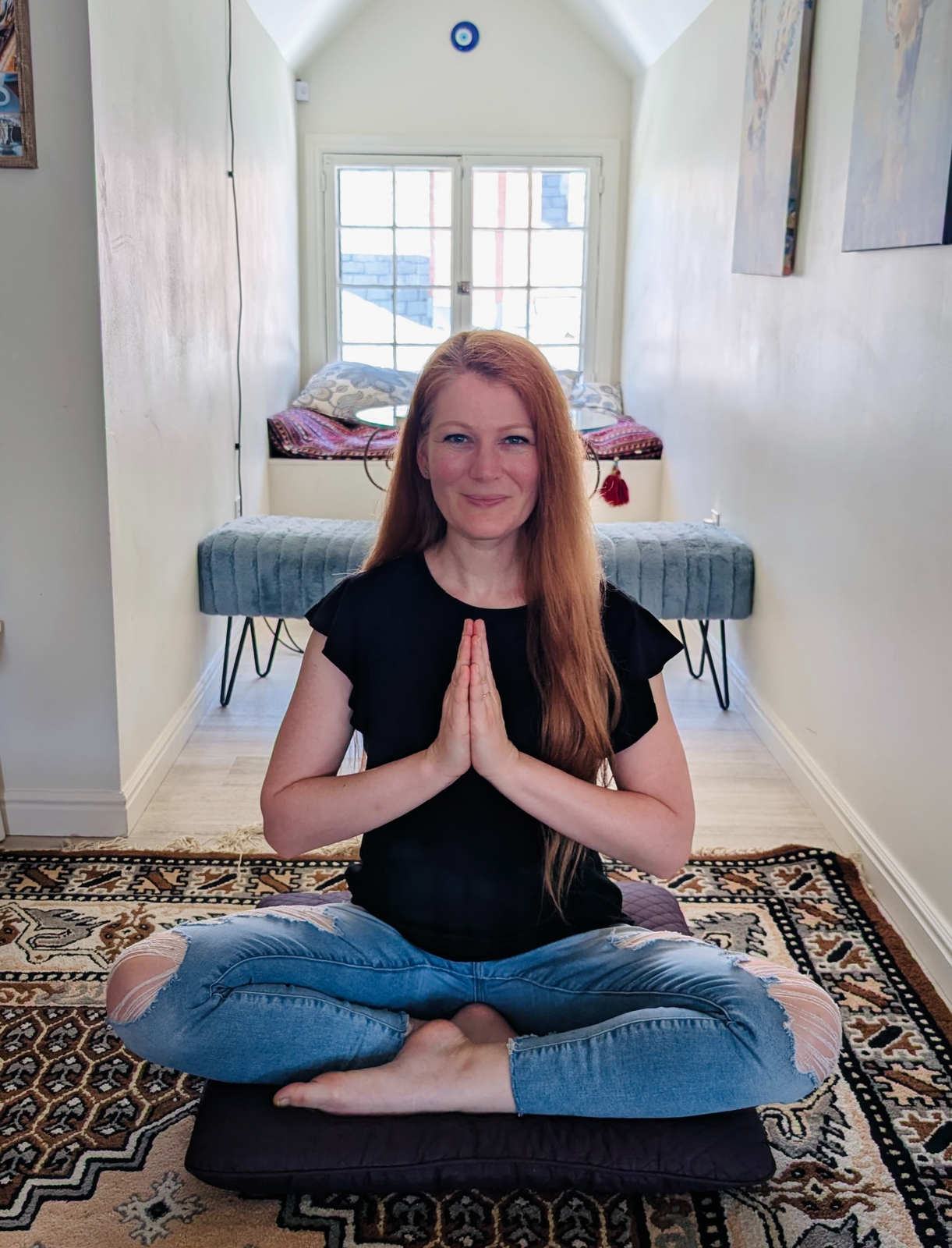 Smiling woman with light red hair in jeans and a black t-shirt, looking confident, in seated yoga pose