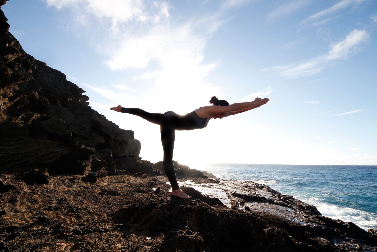 Jennifer balanced in Standing Stick pose on rocks above ocean.