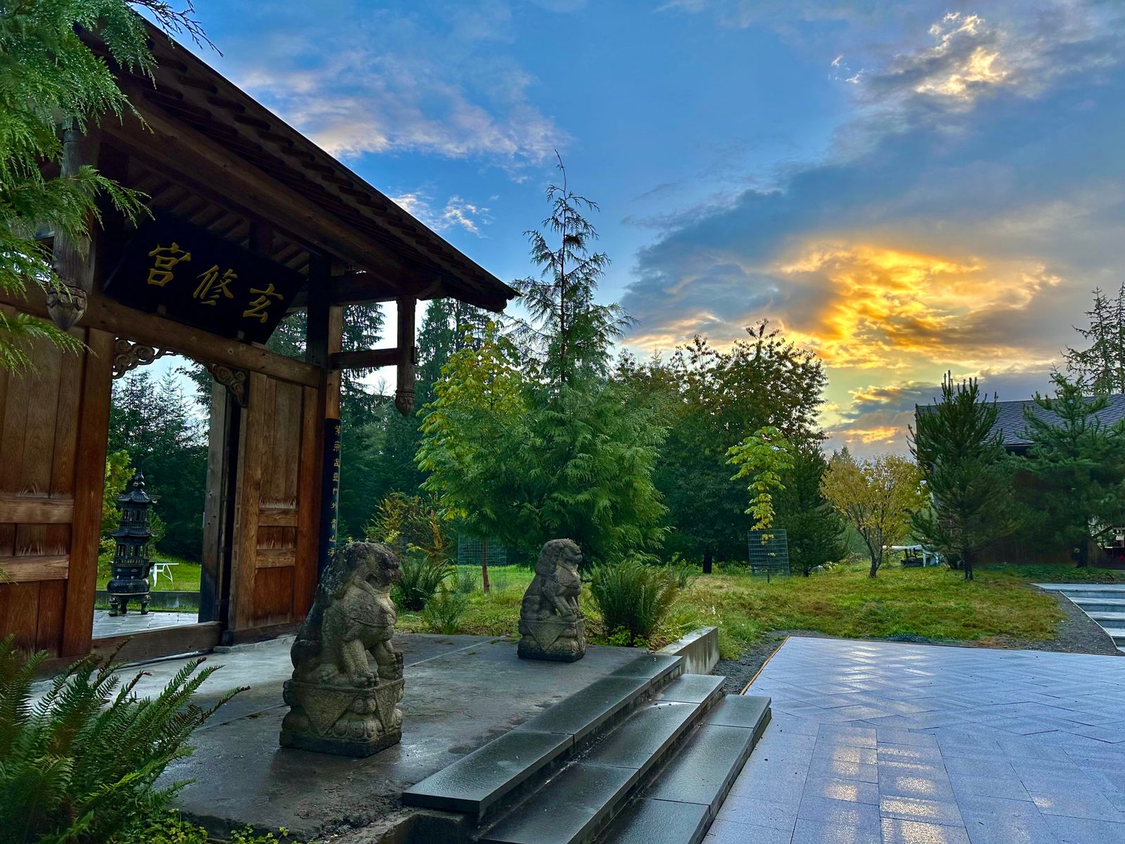 Entrance gate to the Daoist Temple at Xuan Xiu Gong Sanctuary. A tall wooden archway with the Chinese characters for Xuan Xiu Gong above it. Gate is flanked by two stone lions. In the background, a sunset. The perfect place for qi gong, tai chi, and meditation practice during frequent Taoist retreats.