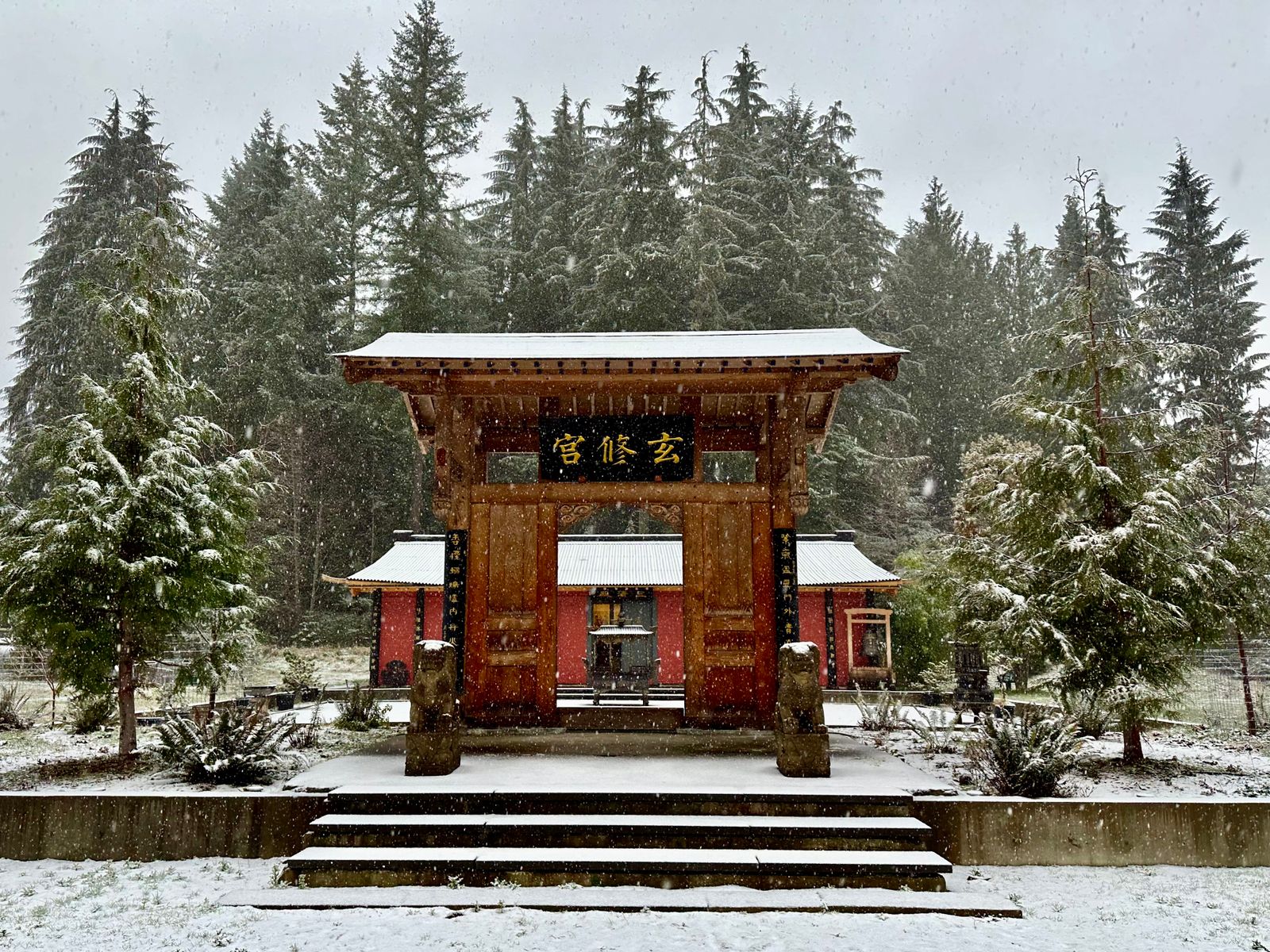 Snow dusts the great gate and ground entering into the Daoist Temple's inner courtyard at Xuan Xiu Gong Taoist Sanctuary. The Taoist temple, itself, is in the background. Evergreen trees present a vibrant backdrop for Daoist cultivation and practice, including qi gong and tai chi.