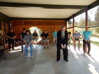 Students practicing qi gong (Daoist movement meditation) with Taoist Master Mo ZhiCheng outside in the moon pavilion at Xuan Xiu Gong Daoist Sanctuary in Snohomish, Washington, USA. Sunlight streams through the round gate and past the evergreen trees beyond the practice space.