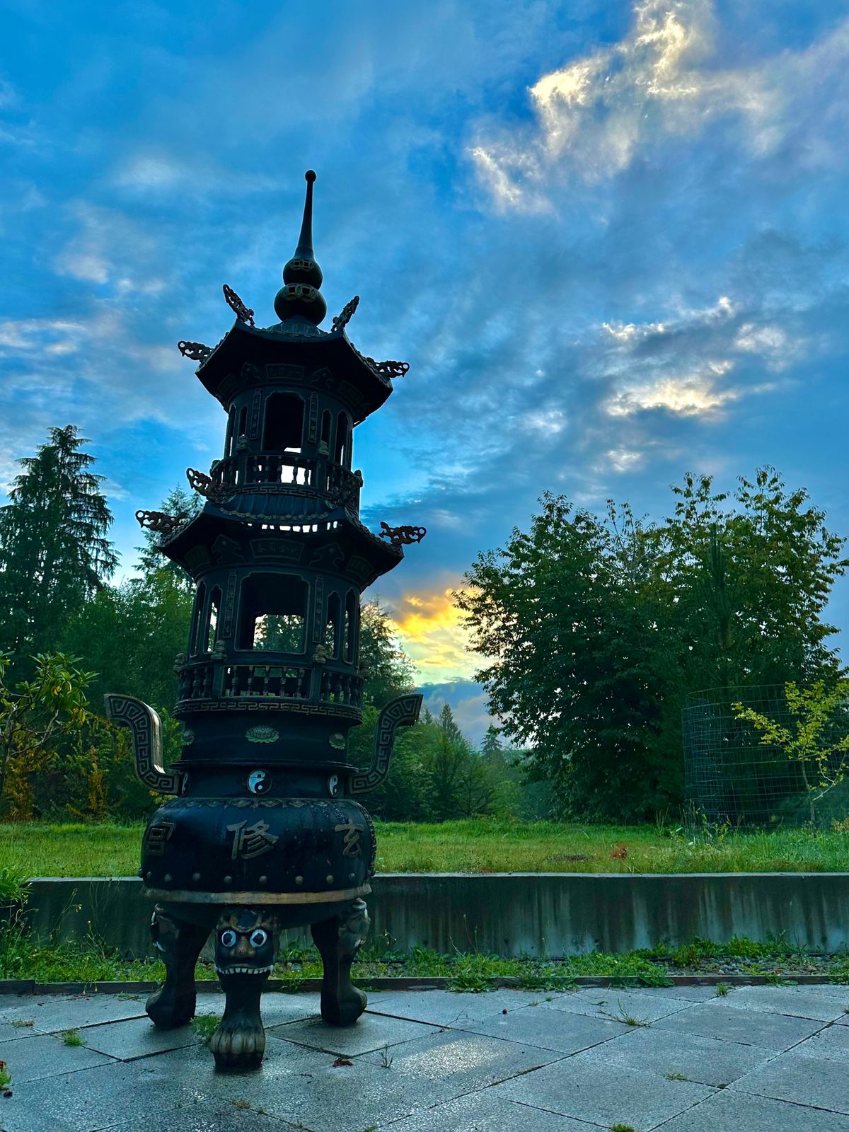 Sunrise above the mountains to the east of the Daoist Sanctuary, Xuan Xiu Gong, casts a golden glow beside an ornate, towering incense burner. Sunrise over the Daoist temple courtyard and retreat center at Xuan Xiu Gong monastery. Entrance gate and incense burners are lit by the orange light of dawn.  The perfect place for qi gong, tai chi, and meditation practice during frequent Taoist retreats.
