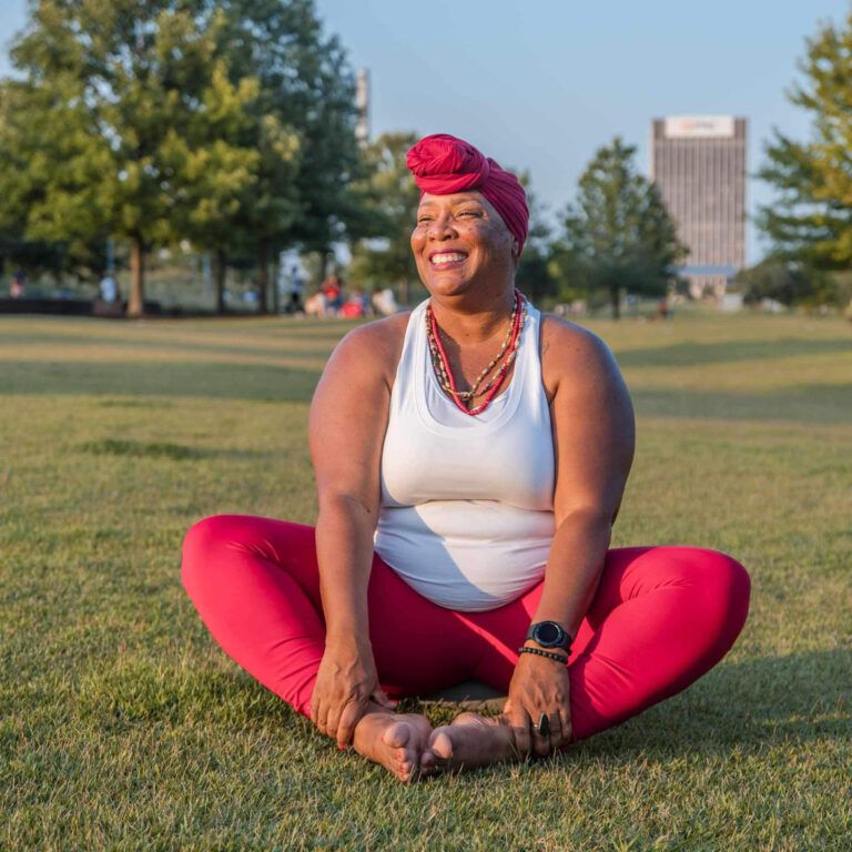A black woman wearing a red head wrap sits in bound angle pose in the park. She smiles widely at the camera.